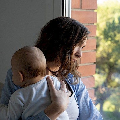 Eine junge Mutter schaut traurig aus dem Fenster. Sie hat ein Baby auf dem Arm