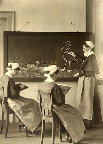 1920 – Ausbildung von jungen Frauen zu Kinderpflegerinnen Eine historische Aufnahme von 1920 in schwarz-weiß zeigt die Ausbildung von jungen Frauen zu Kinderpflegerinnen in Großheppach. Schülerinnen sitzen im Klassenzimmer vor einer Tafel.